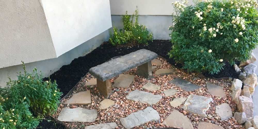 Stone bench in a small garden bed with stone and gravel walkway, and lush green plants.