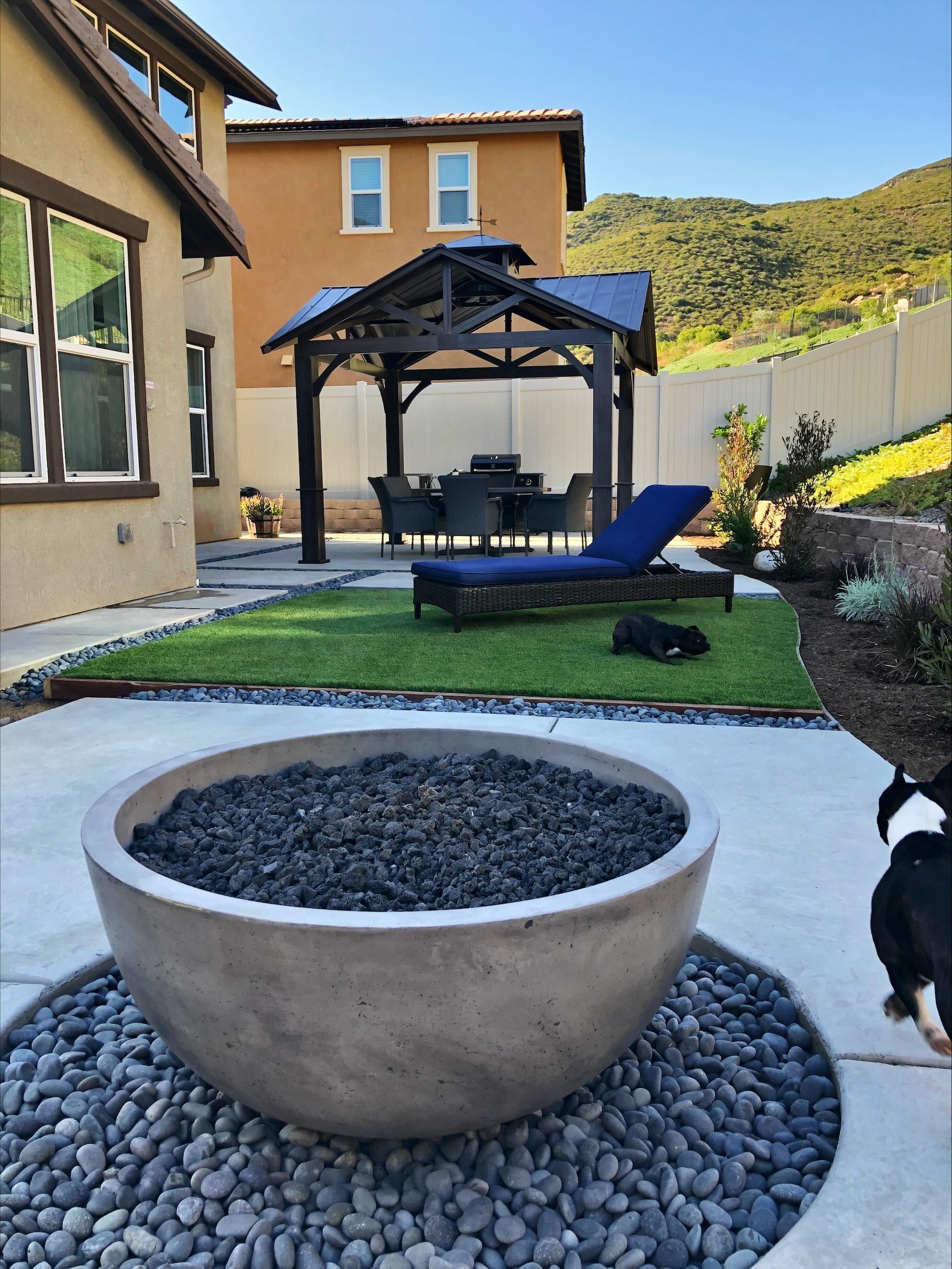 Backyard with a gazebo, lounge chair, fire pit, and a dog. Sunny day, green grass, and mountains in the background.