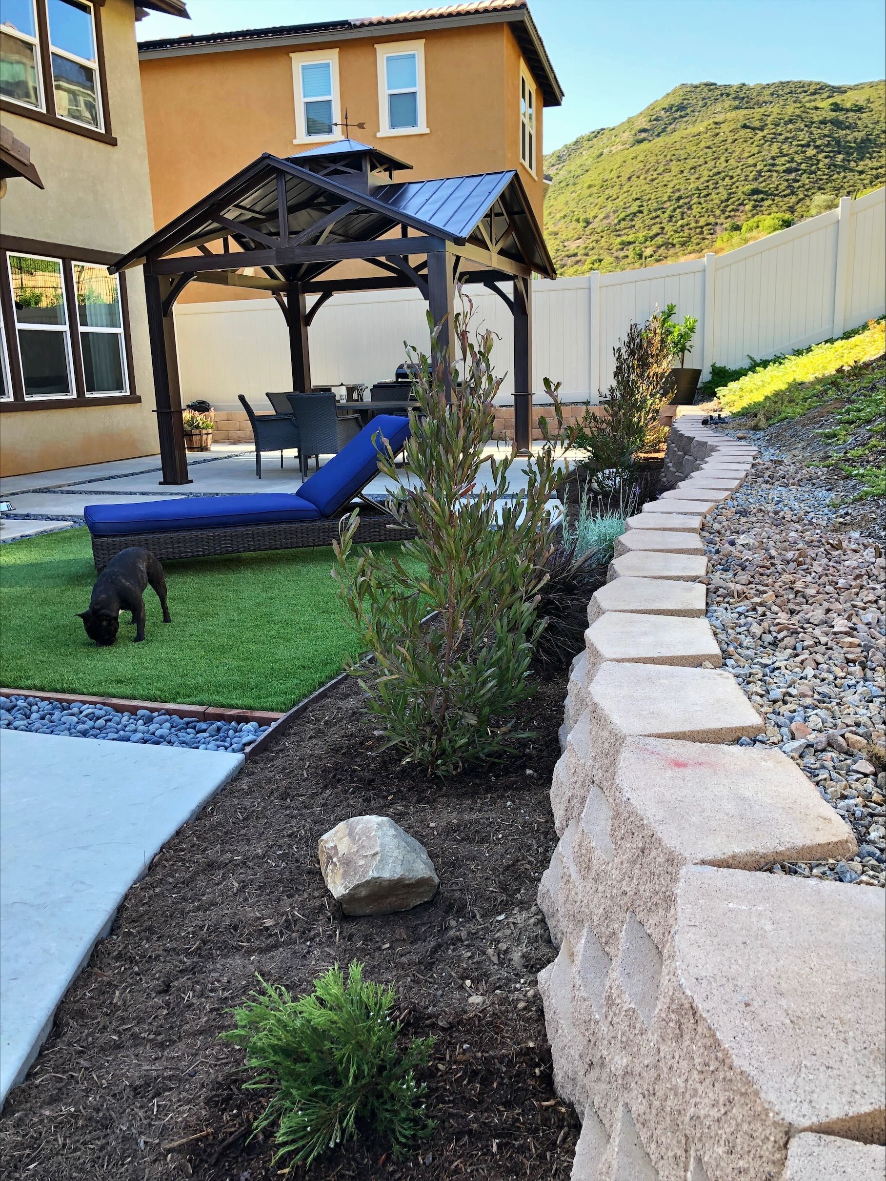 Backyard scene with a gazebo, lounge chair, stone pathway, and a dog.
