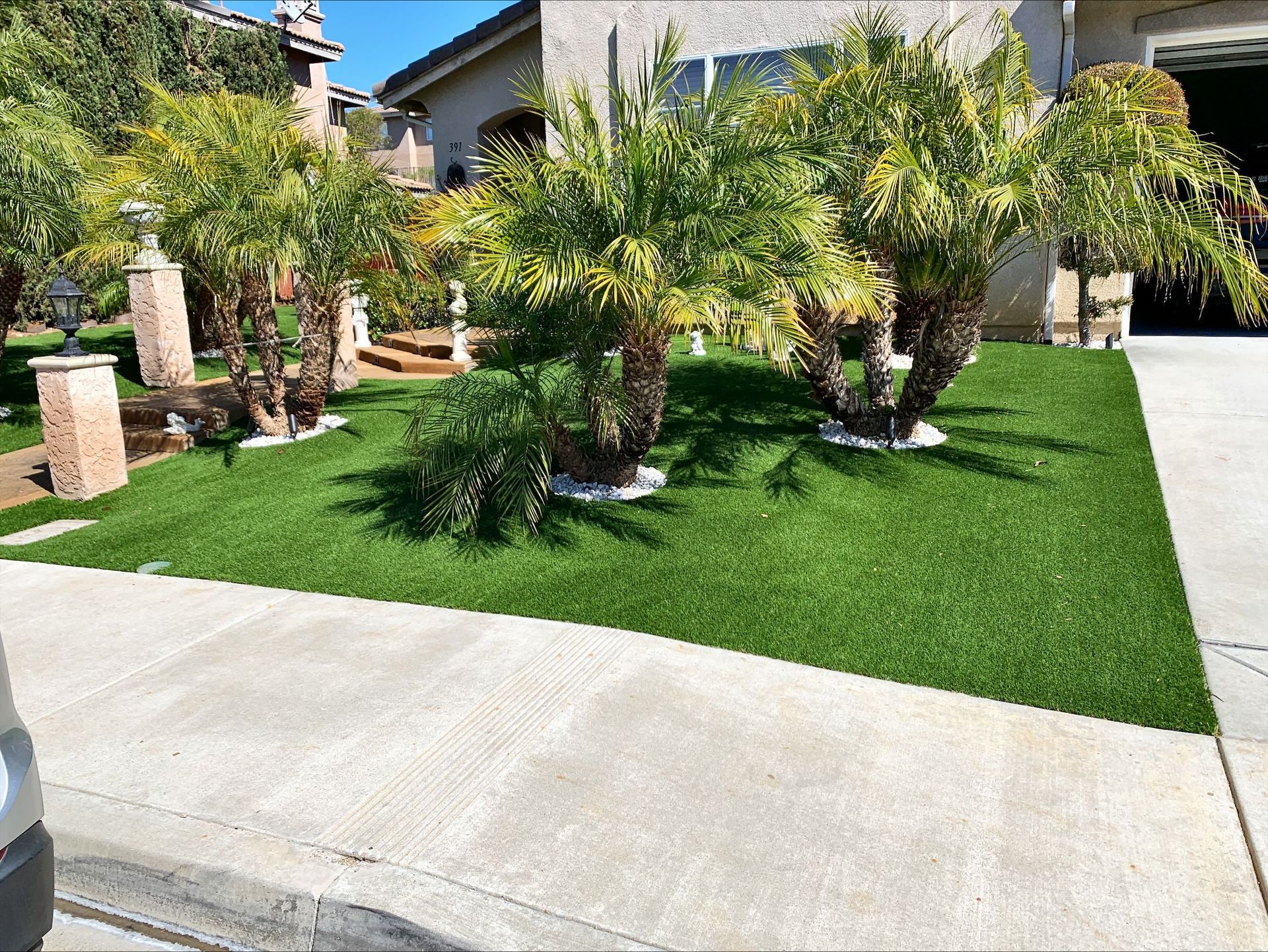 Lush green artificial turf in front yard with palm trees and a concrete sidewalk.