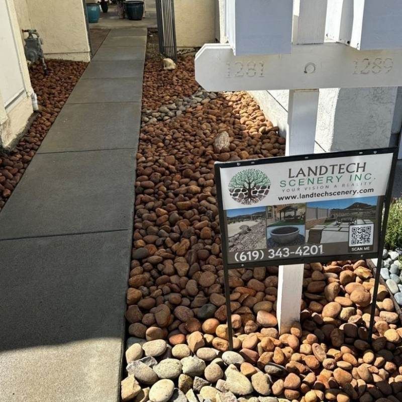 A concrete walkway next to a bed of brown river rocks. A white mailbox and a Landtech Scenery sign are present.