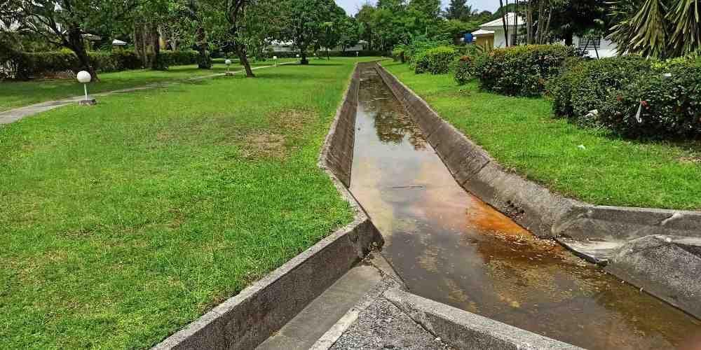 A concrete waterway runs through a grassy park, trees and bushes in the background, cloudy day.