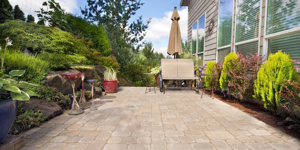 Sunny backyard patio with paver path, lounge chair, umbrella, and lush green garden border