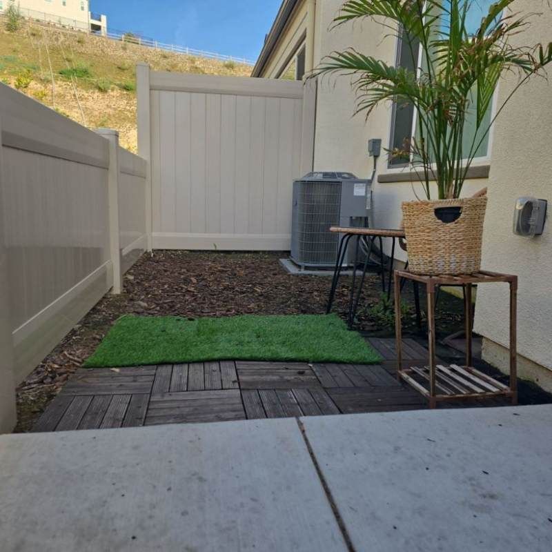 A small outdoor space with a wooden patio, artificial grass, and a potted palm. A/C unit and white fence present.