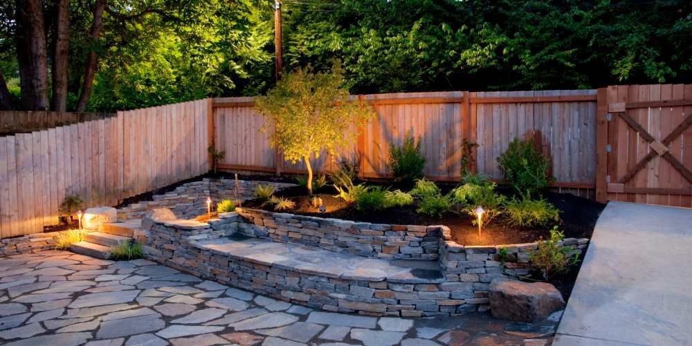 Backyard patio with stone steps, landscaping, and lit trees, beside wooden fences.
