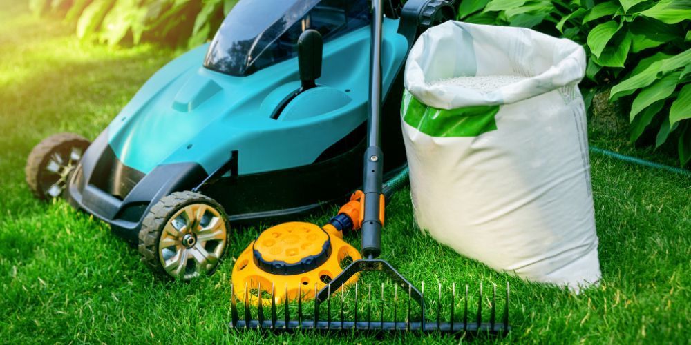 Lawn mower, bag of fertilizer, rake, and yellow sprinkler on a green lawn.