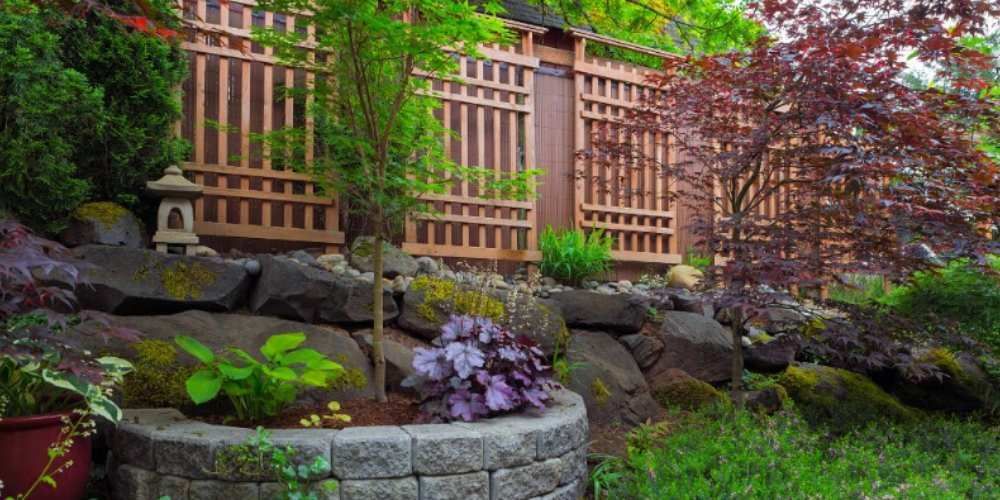 Japanese garden with a wooden lattice fence, stone walls, and lush green and purple foliage.