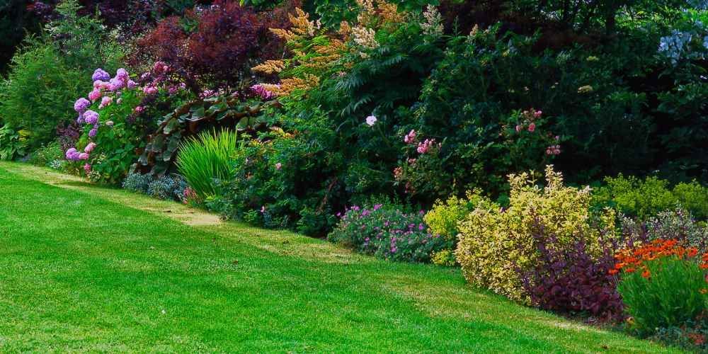 Lush green lawn slopes down to a diverse garden bed filled with colorful flowers and shrubs.
