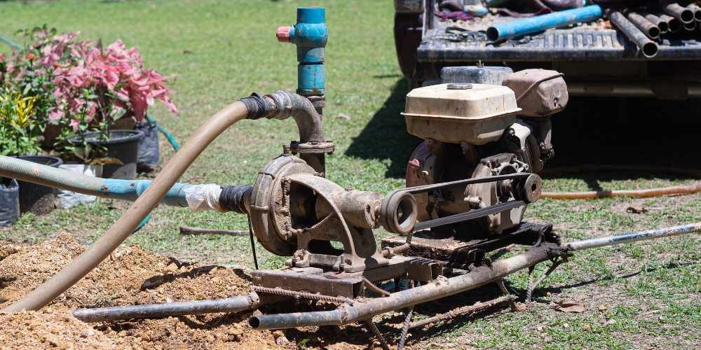 Old portable water pump with hoses on grass near potted flowers and a utility truck.