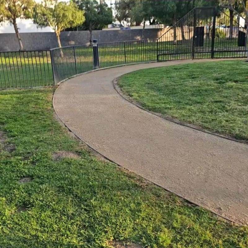Curving paved path through a grassy park, bordered by a black metal fence.