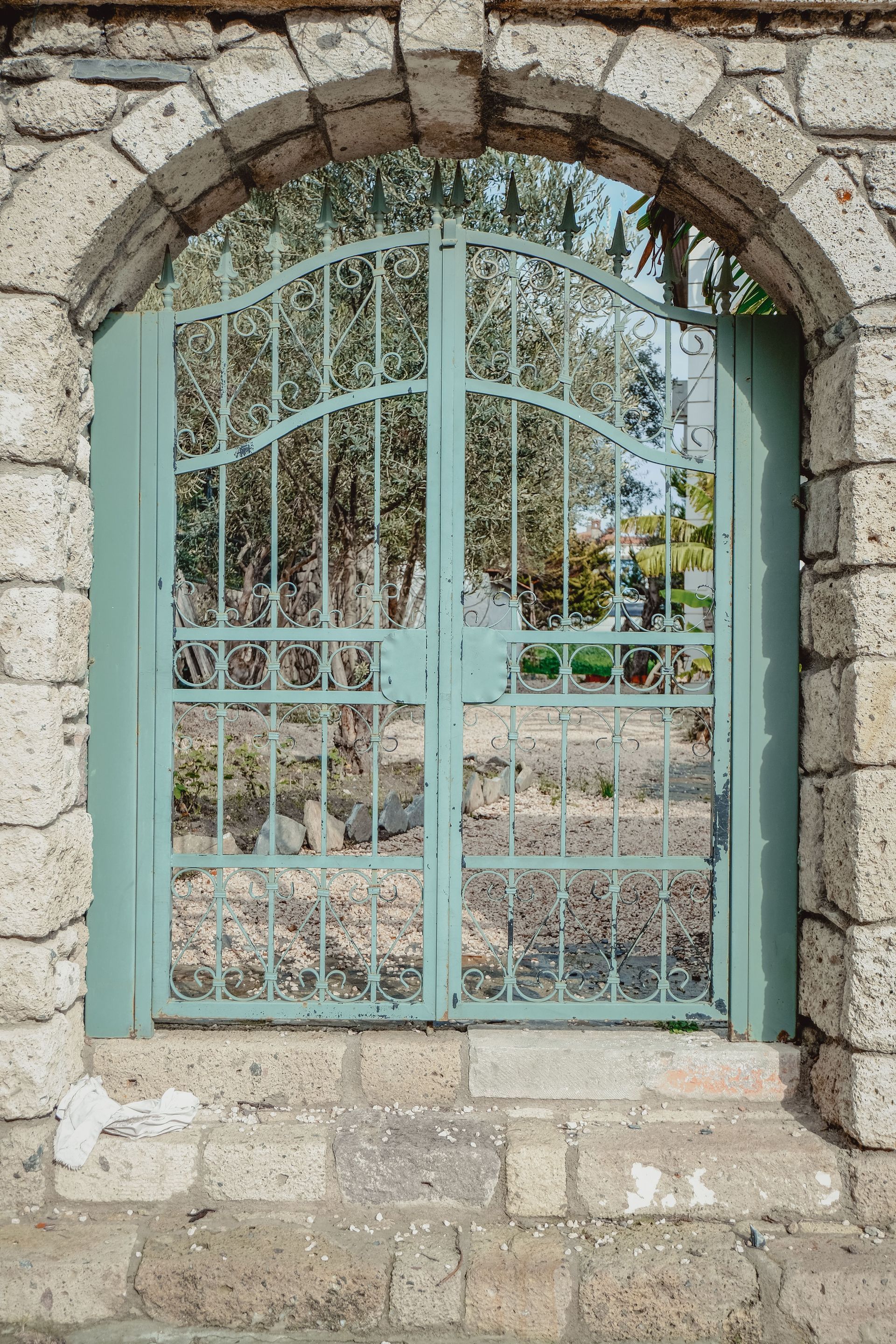 Stone arch framing a pale blue metal gate with reflections of trees behind it.