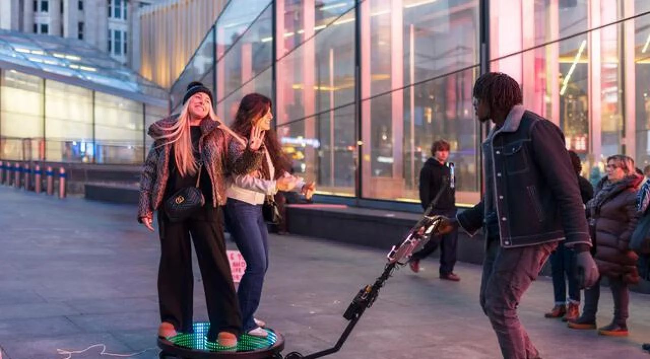Two women on a hoverboard-like device on a city street, with a man nearby and glass buildings in the background.