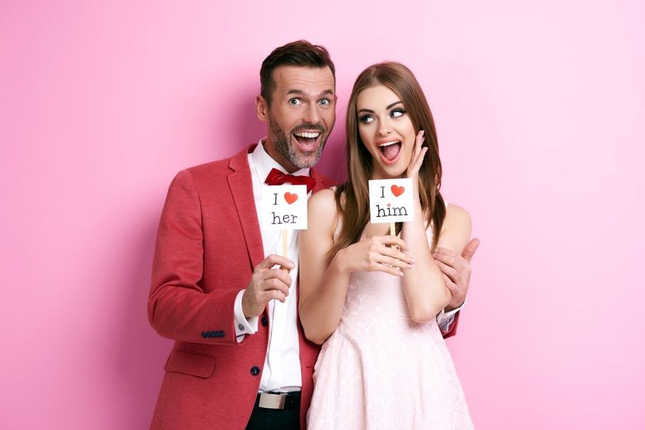 Two smiling people in pink outfits holding small cards against a pink background