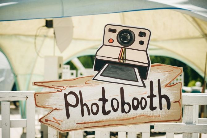 Hand-painted “Photobooth” sign with a cartoon camera on a white fence outdoors