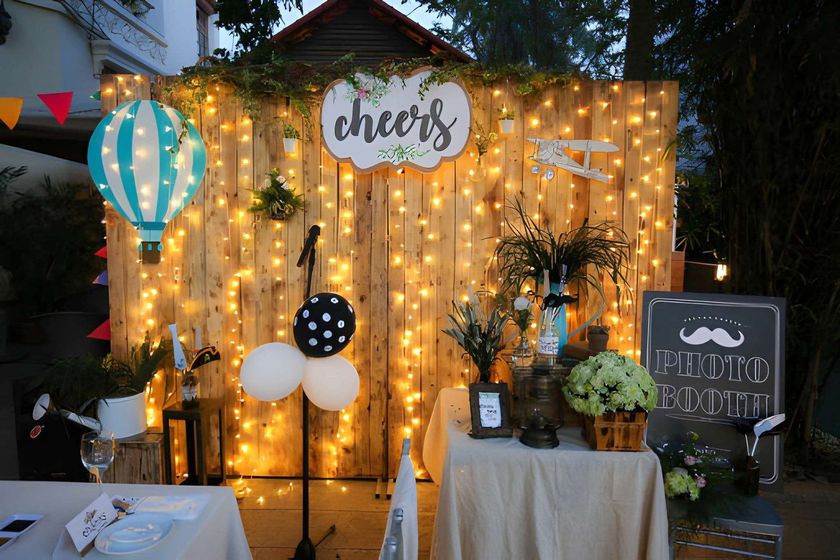 Decorated outdoor party backdrop with string lights, balloons, and a “cheers” sign under a canopy.