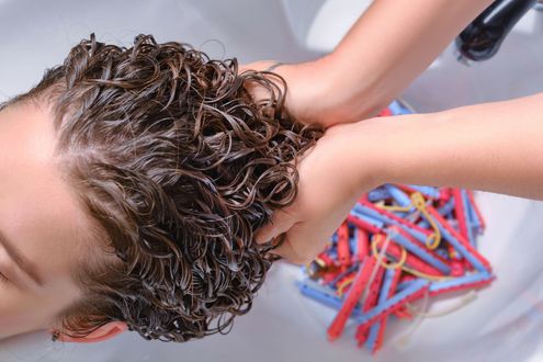 Hair being washed at a sink, with hands massaging shampoo into the scalp and colorful hair rollers nearby