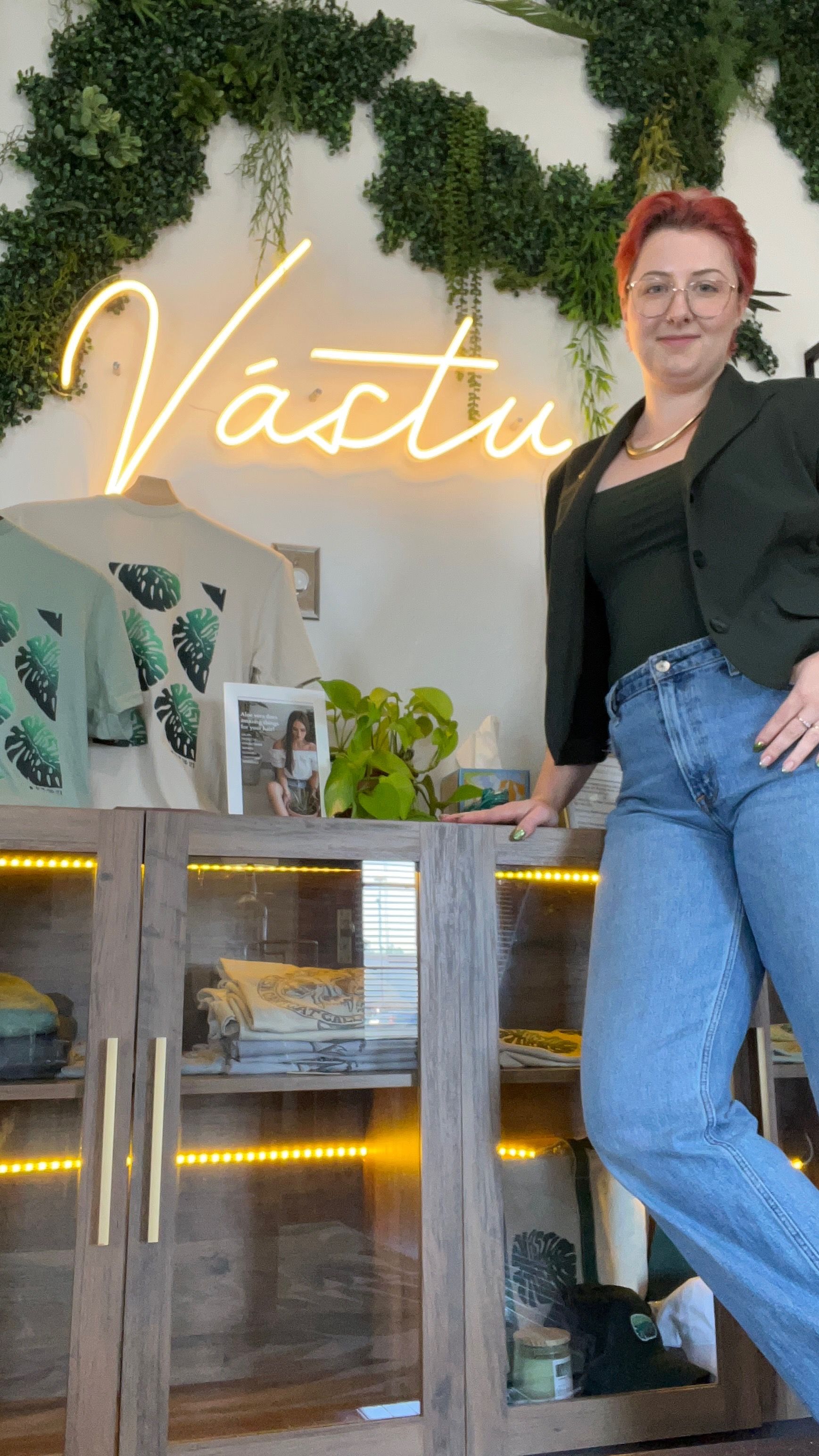 Woman standing by a neon-lit bar with shelves and plants, wearing a black blazer and jeans.