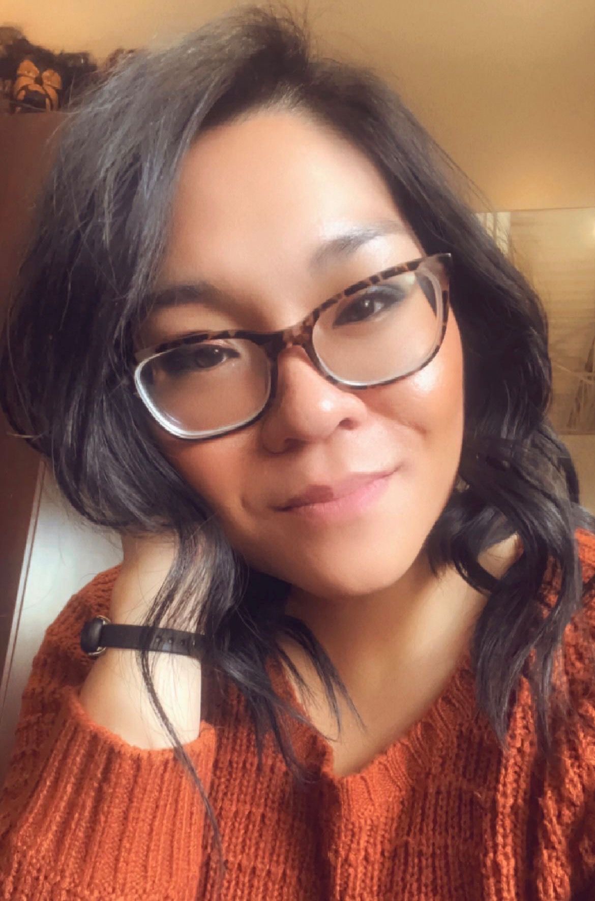 Portrait of a person with dark hair and glasses, wearing an orange sweater and smiling softly indoors