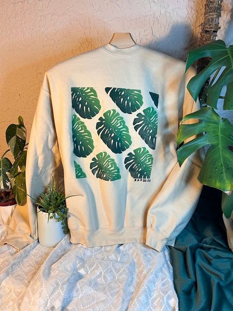 Cream sweatshirt with green leaf print displayed on a draped white cloth beside houseplants