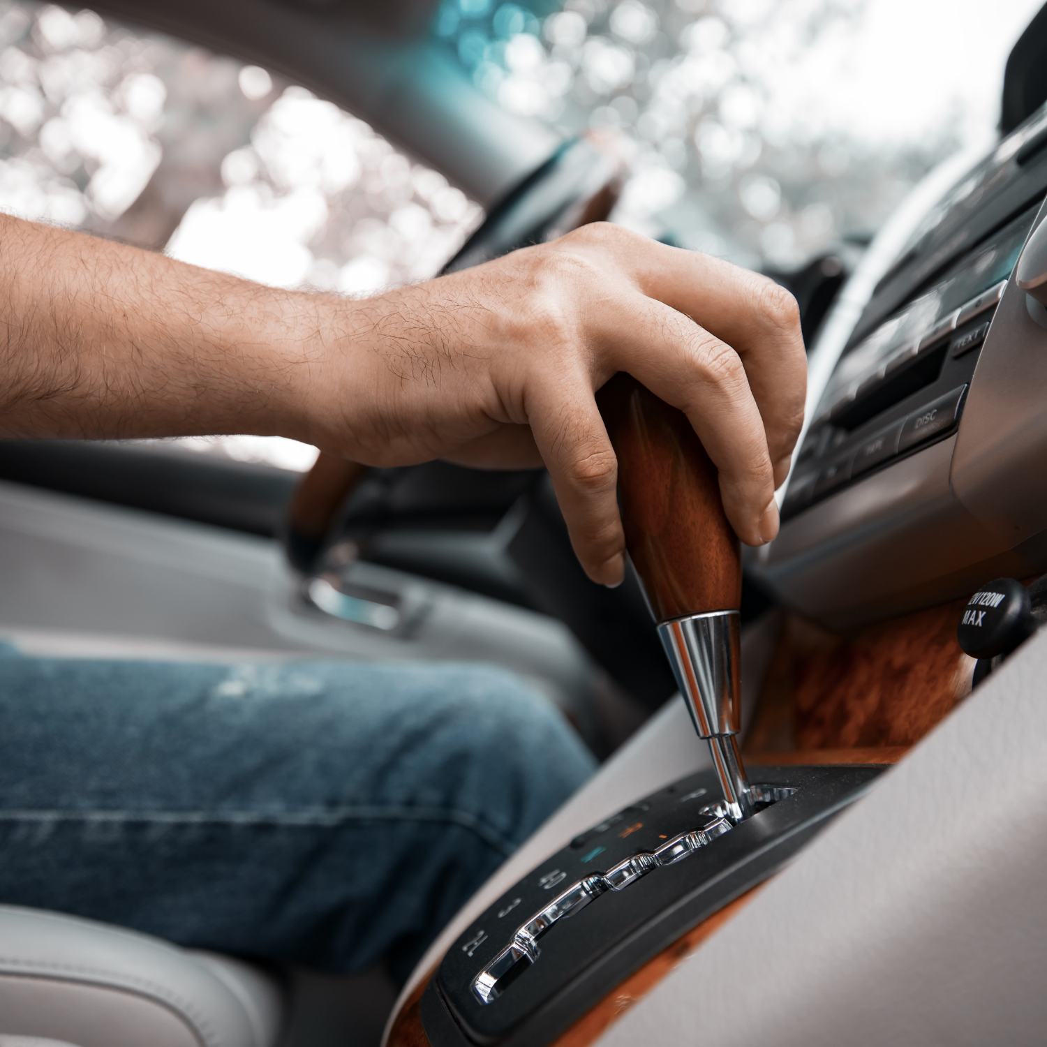 Hand on wooden gear shift in a car interior, with blue jeans and dashboard visible