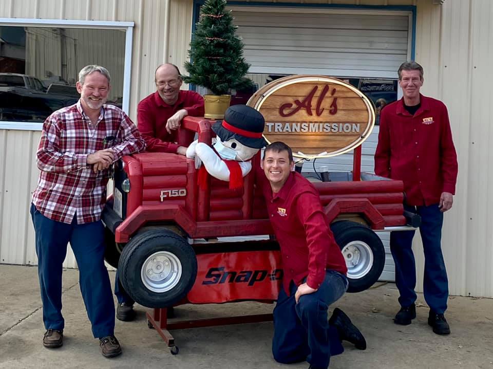 Four men pose with a red toy truck and snowman in front of an All’s transmission mission sign.
