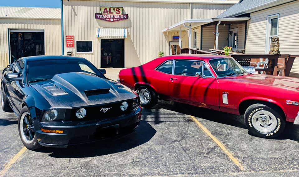 Two classic muscle cars, a black Mustang and red coupe, parked outside a building with an AOS sign.