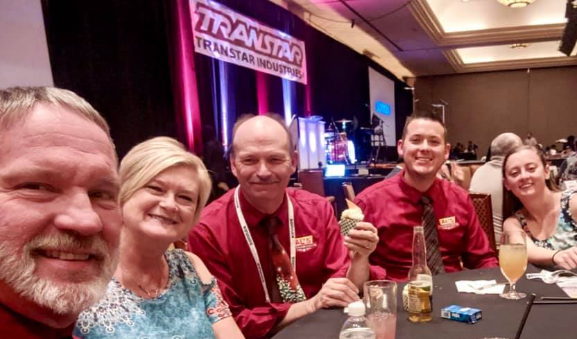 Four smiling people seated at a conference table with drinks, in front of a stage with a TRANSNAT banner.