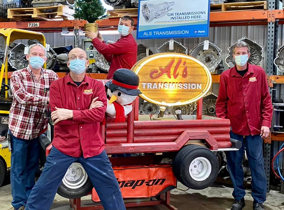 Four masked workers posing in a garage beside a red cart labeled “AT Transmission”