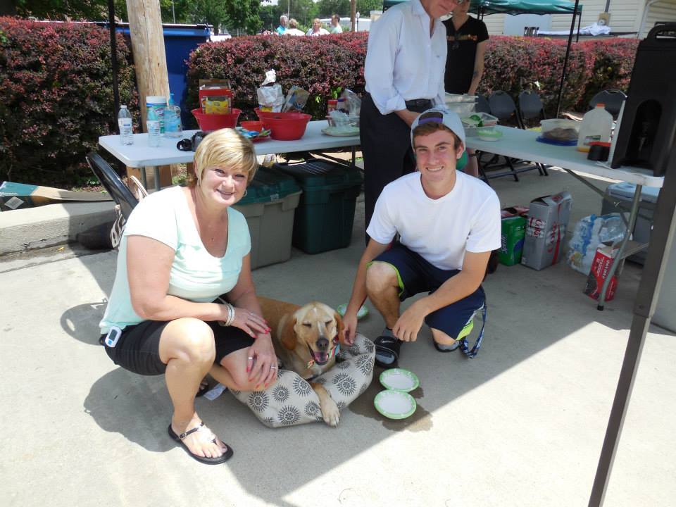 Two people crouch beside a small dog at an outdoor market stall with potted plants and pet bowls