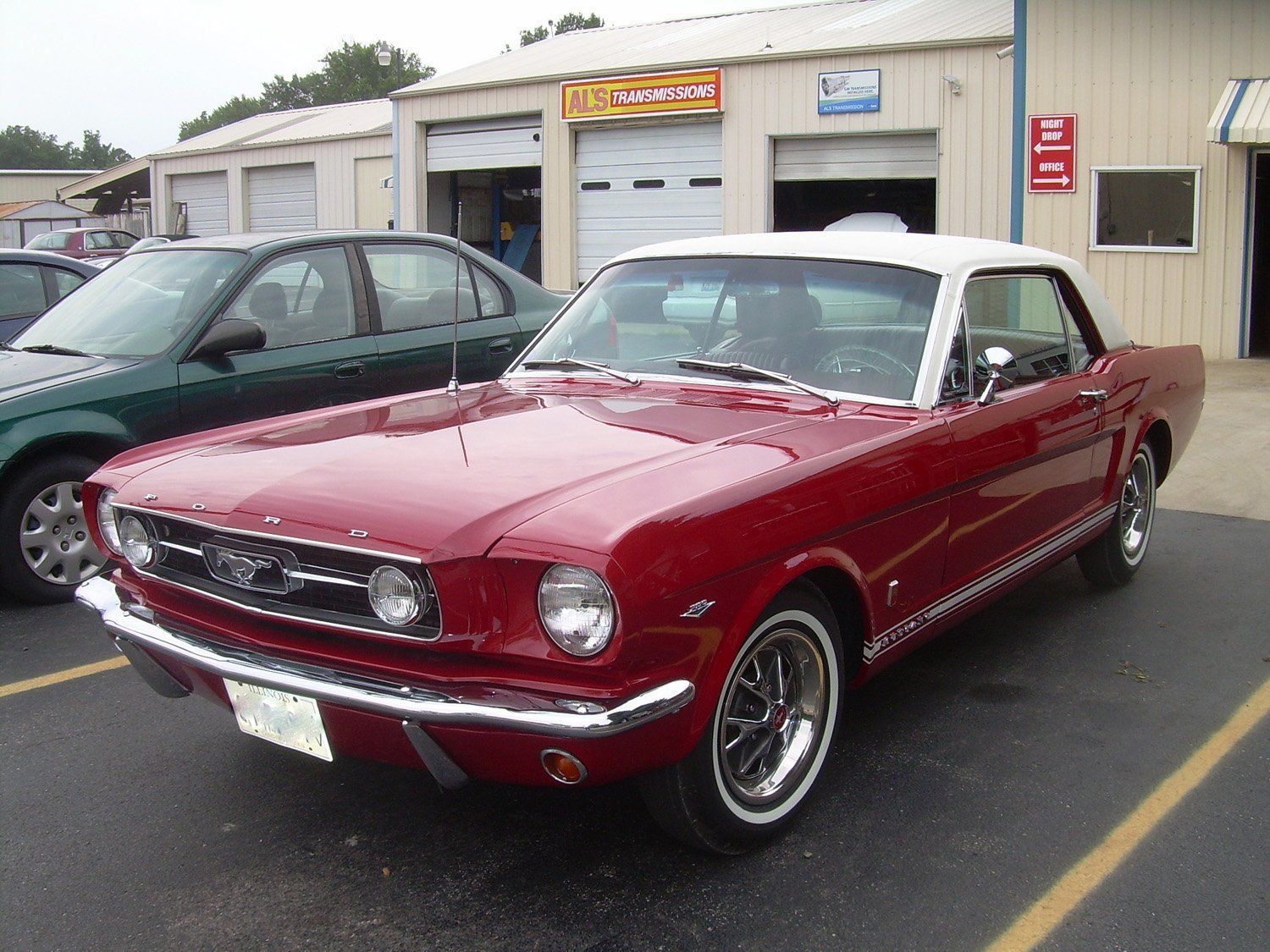 Red classic Mustang convertible parked outside a garage, with a green car beside it.