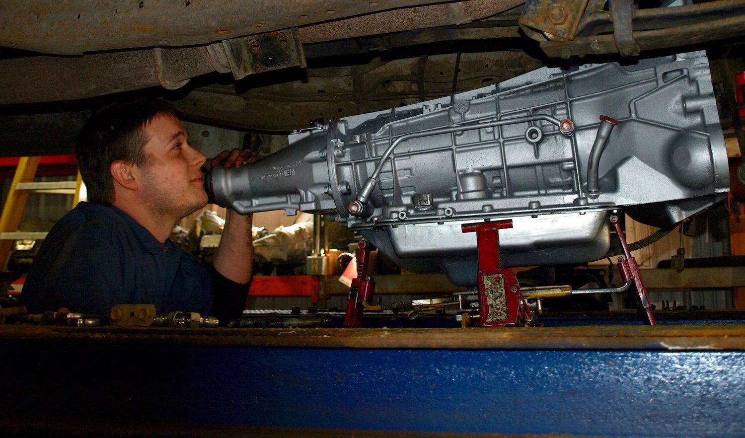 Mechanic inspecting a large silver transmission under a vehicle on a lift in a garage