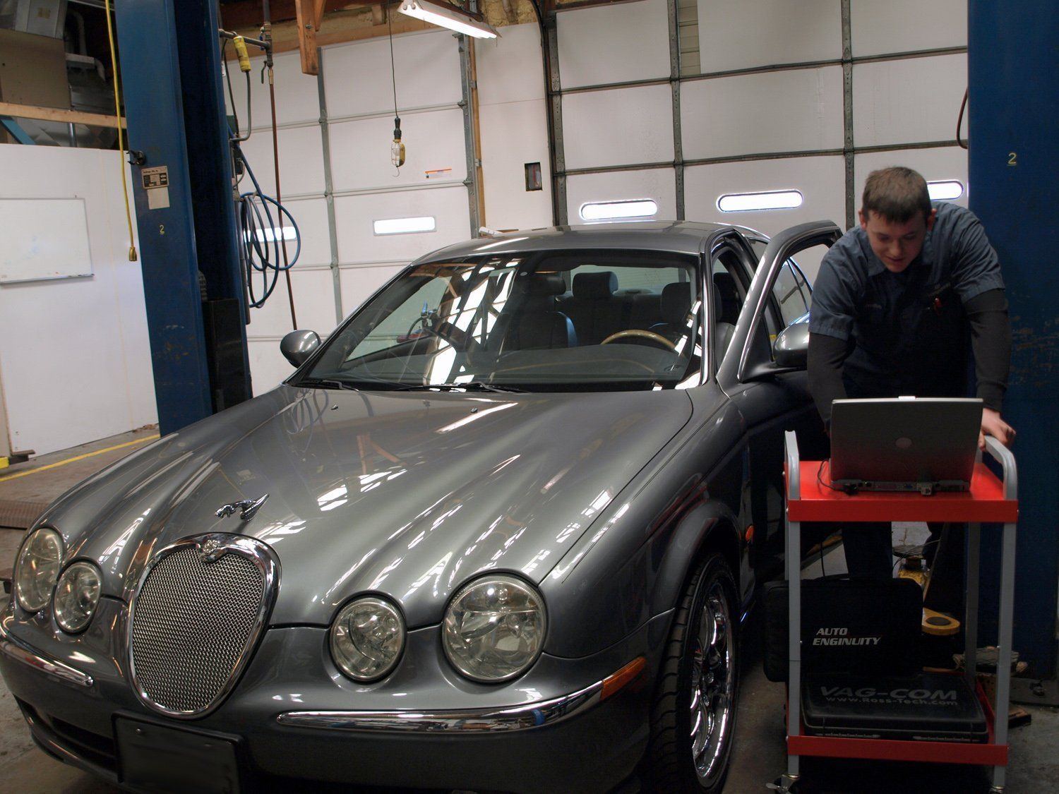 Gray vintage car in a workshop, with a man leaning over a laptop on a red cart.