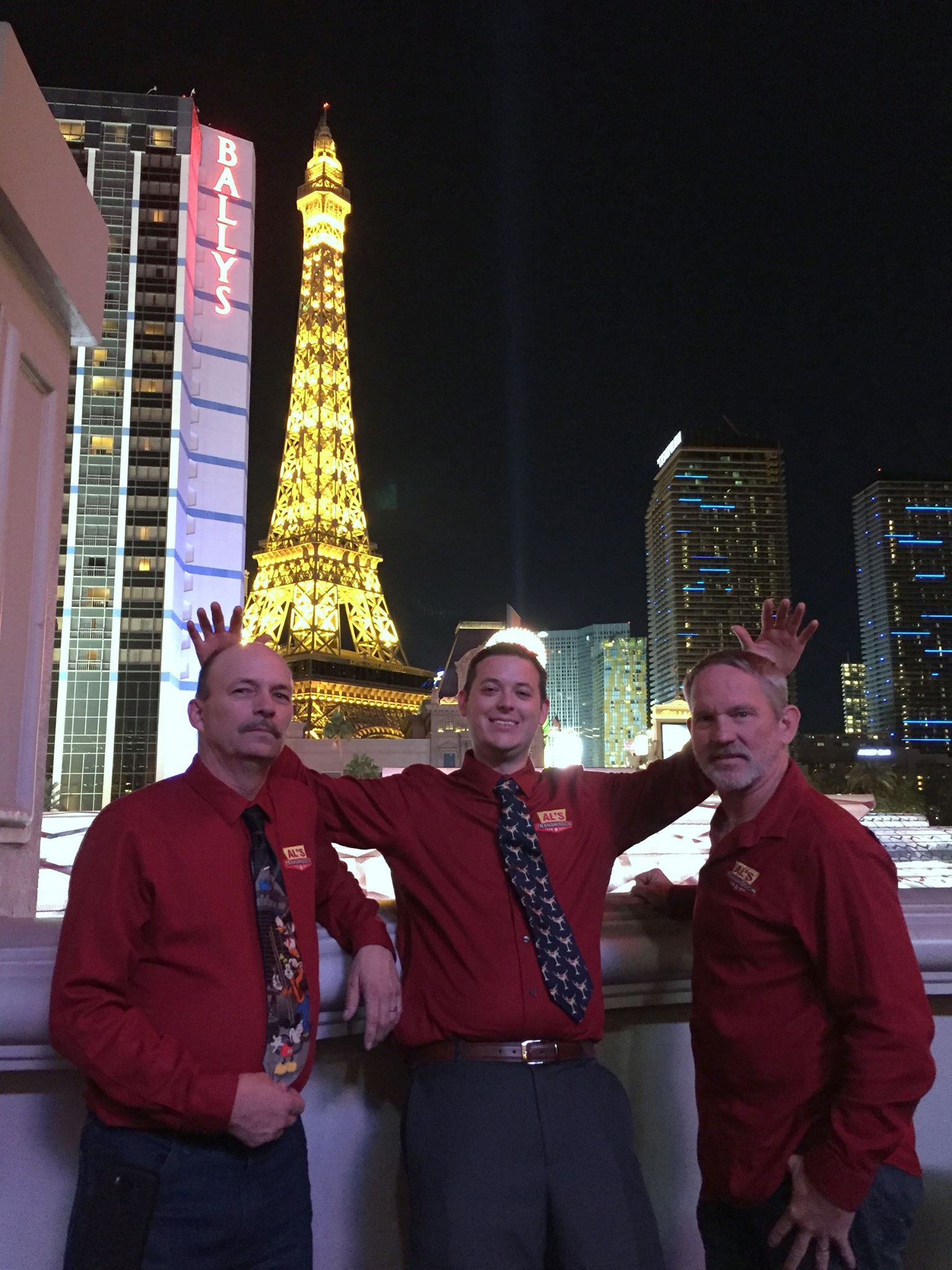 Three men in red shirts pose at night with a lit tower and city buildings behind them.