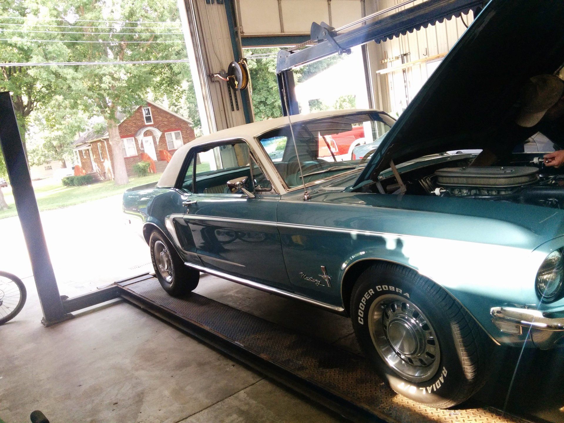 Classic teal convertible on a lift in a garage, hood open, with a red house visible outside.