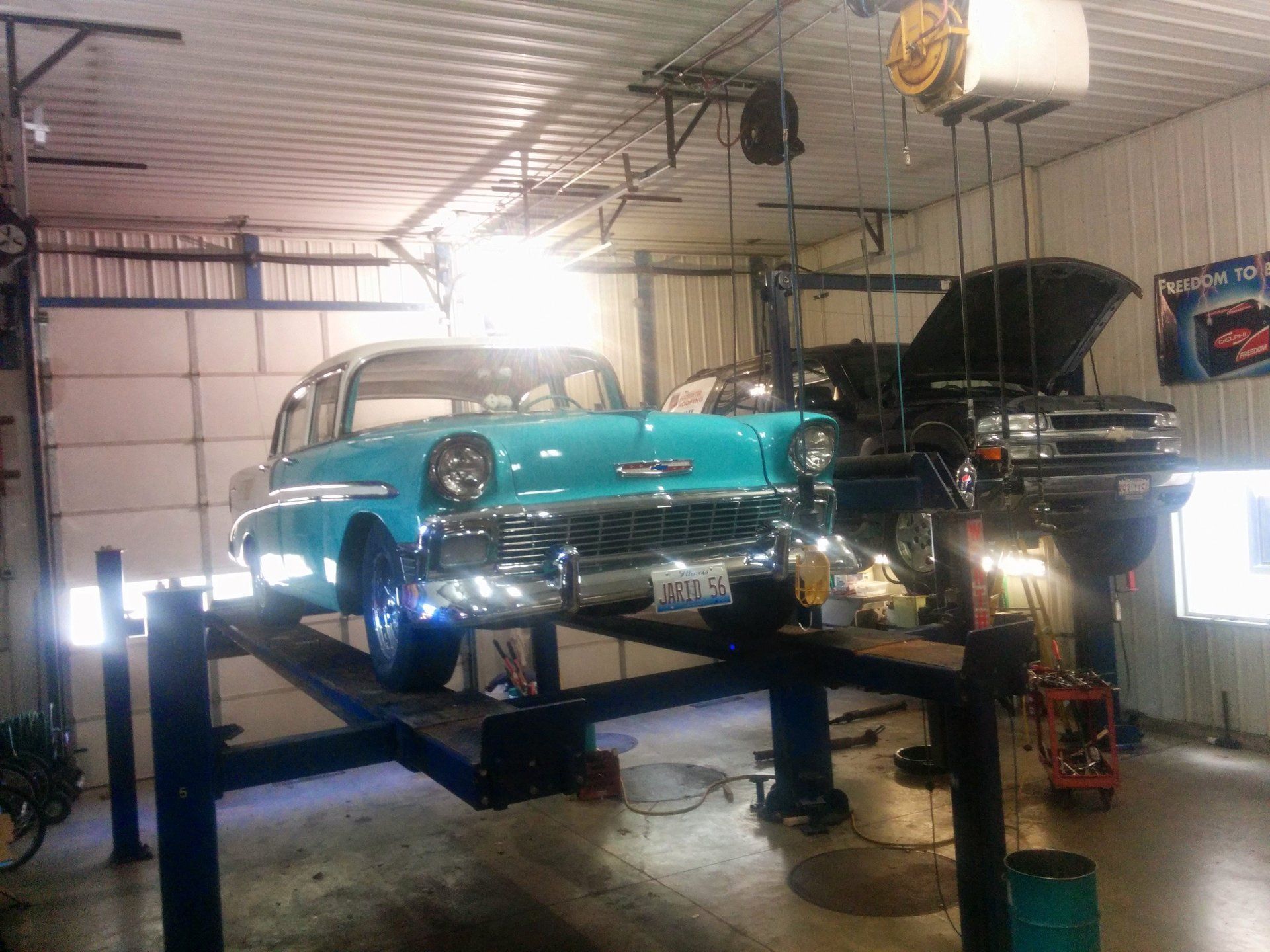 Vintage turquoise car on a lift in a garage, with another car and an open hood nearby.