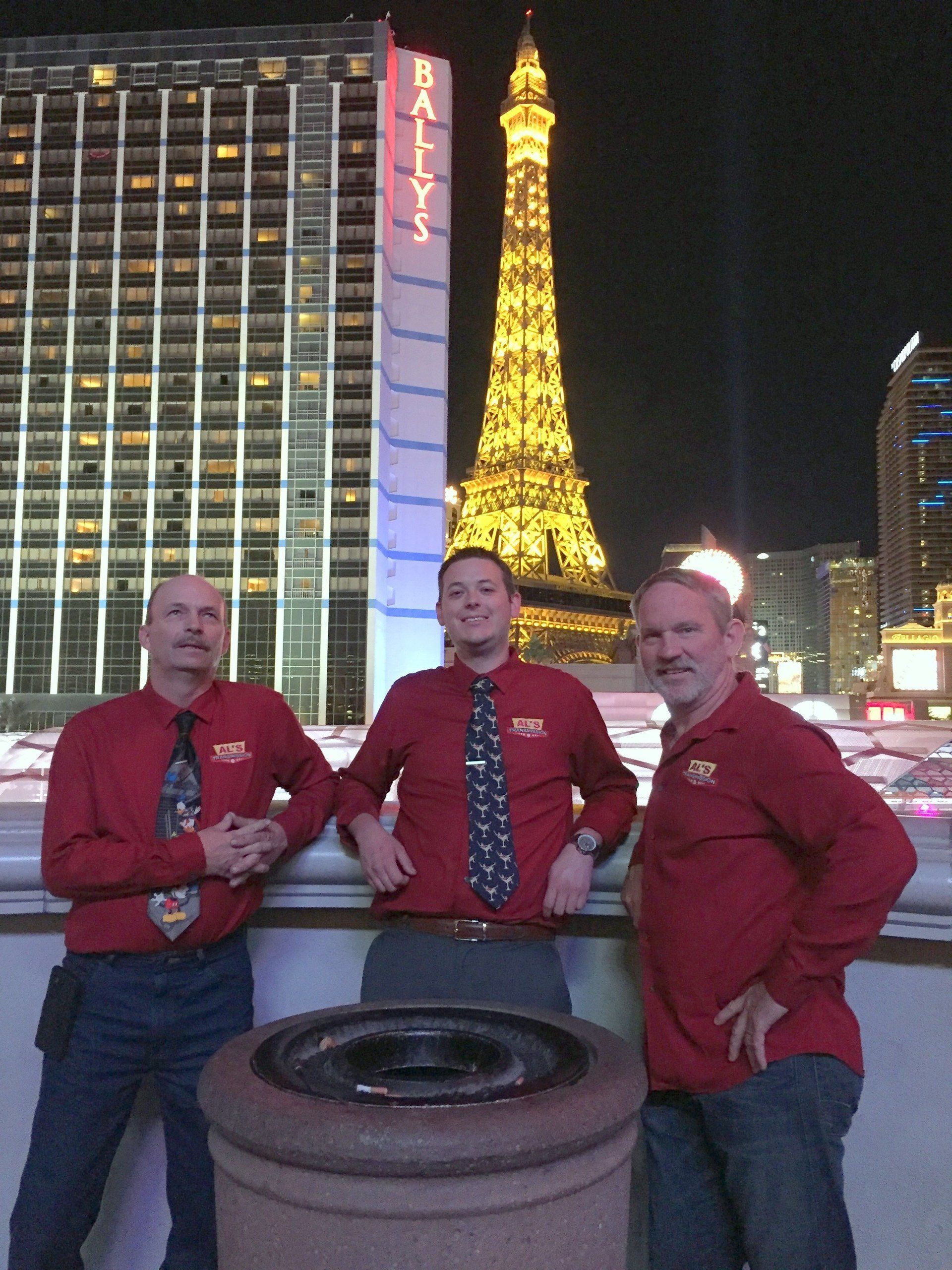 Three people in red shirts pose at a rooftop with the illuminated Eiffel Tower in the background.