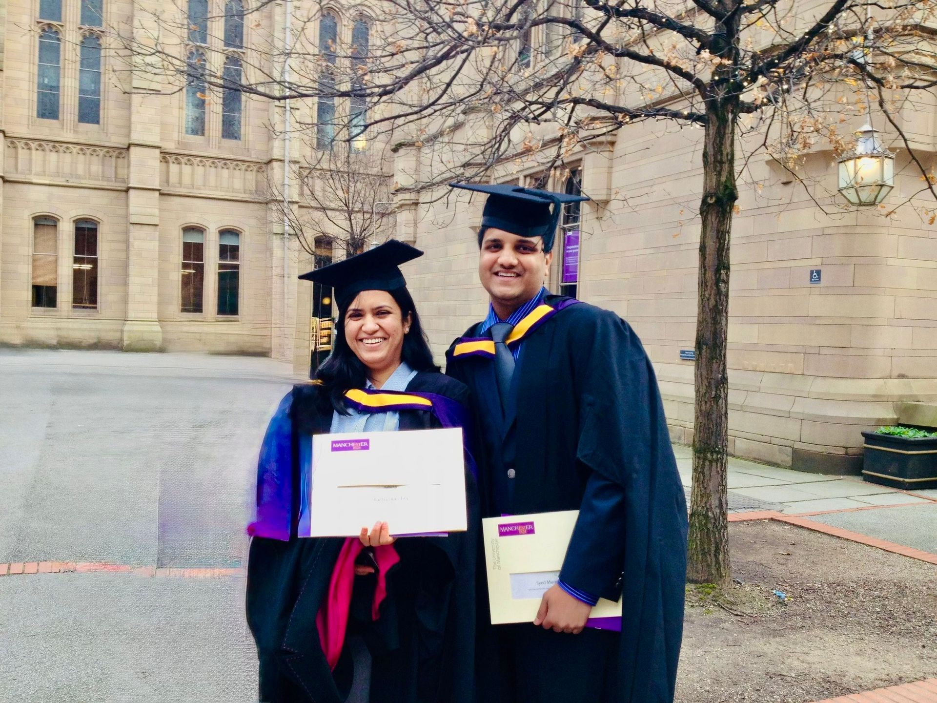 Two graduates in caps and gowns hold diplomas, smiling in front of a stone building.
