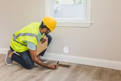 Worker in a yellow safety vest and hard hat cleaning a floor vent in a room with wood flooring.