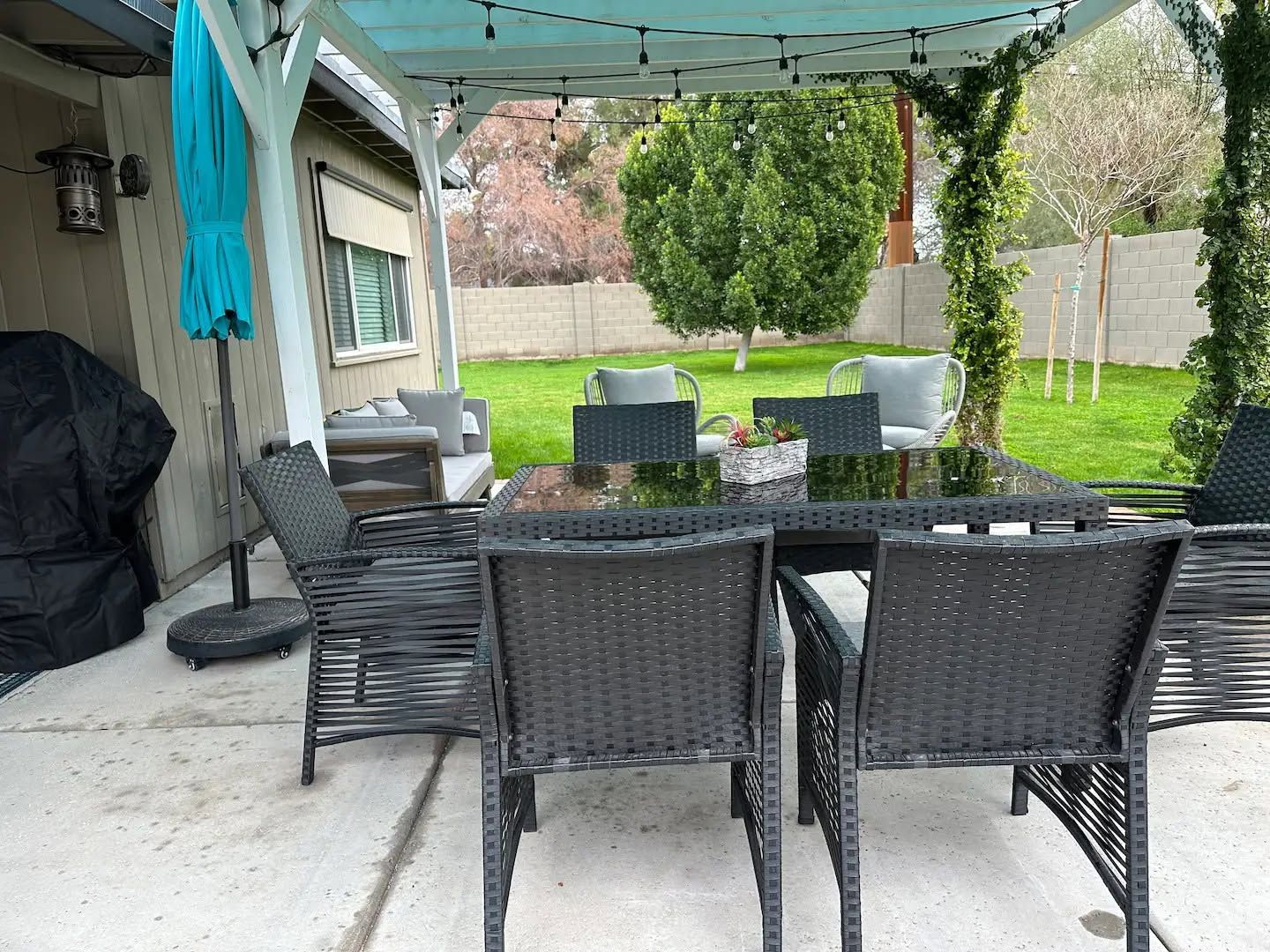Covered patio with black wicker dining set, green lawn, and backyard chairs under a blue canopy