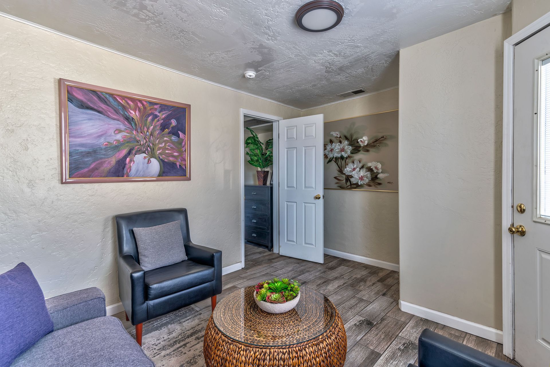 Living room with black leather chair, gray couch, round table, floral art, and open door.