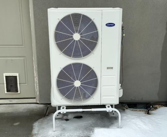 A carrier air conditioner is sitting in the snow in front of a garage door.