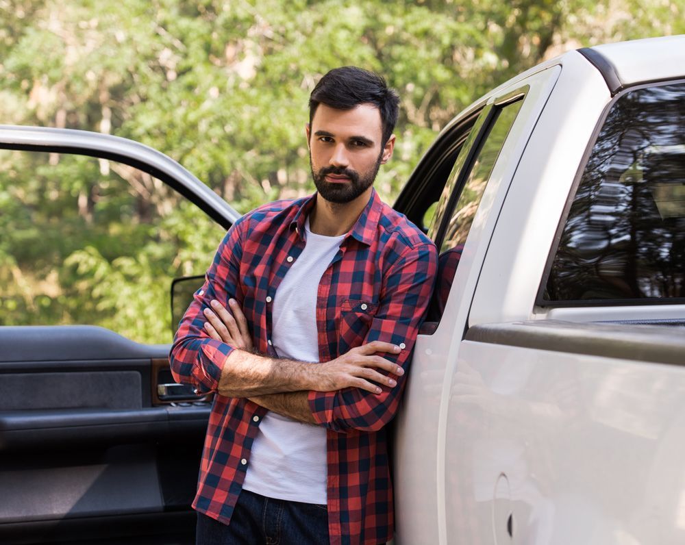 Man leaning against a silver car with arms crossed, beside an open door, wearing a red plaid shirt.