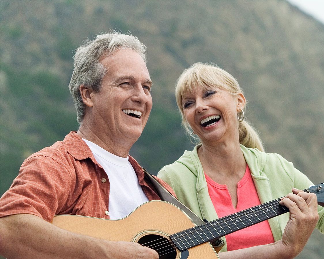 Two smiling people outdoors, one playing an acoustic guitar in front of a mountain backdrop