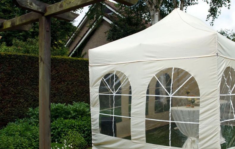 White event tent with arched windows in a wooded backyard beside a house