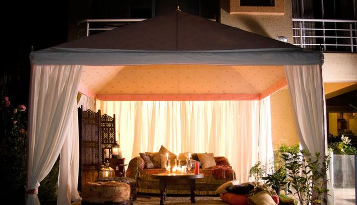 Lit outdoor cabana lounge with sheer curtains, warm lights, and sofa seating at night