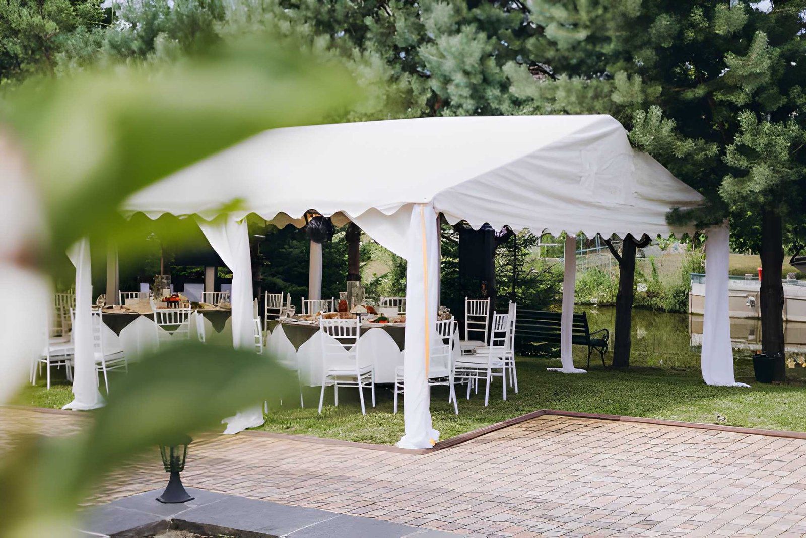White canopy tent with tables and chairs set up outdoors among trees, beside a gravel path.