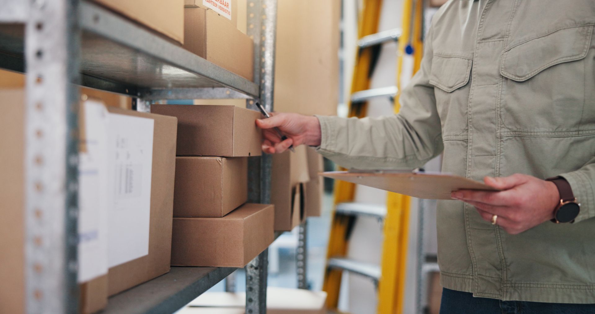 A person checking stacked packages on a shelf with a clipboard in a mail processing area