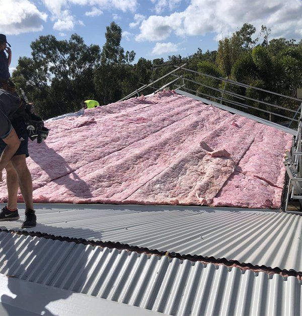 A Man Is Standing On Top Of A Roof Covered In Pink Insulation — Posty’s Roofing In Airlie Beach, QLD
