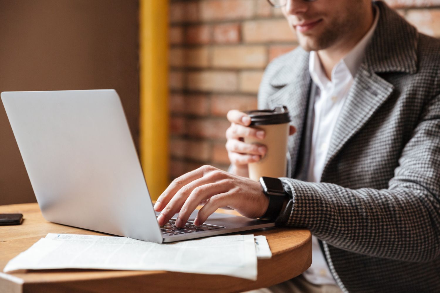 Person working on a laptop at a table while holding a coffee cup in a brick-walled café
