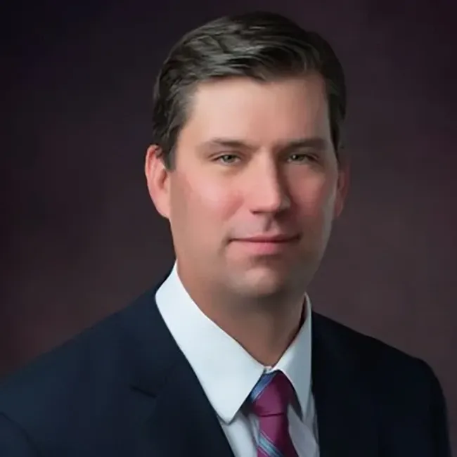 Studio headshot of a man in a dark suit and pink tie against a dark background